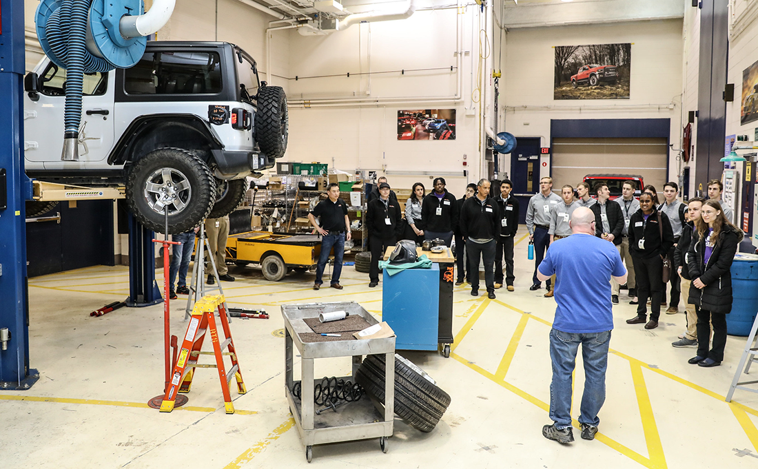 Man speaking to group in garage setting with vehicle suspended for maintenance