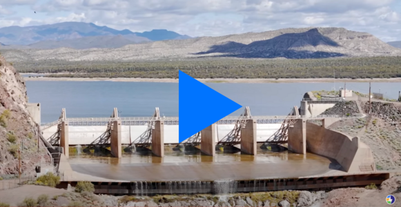 A dam sits in water against a mountain background.