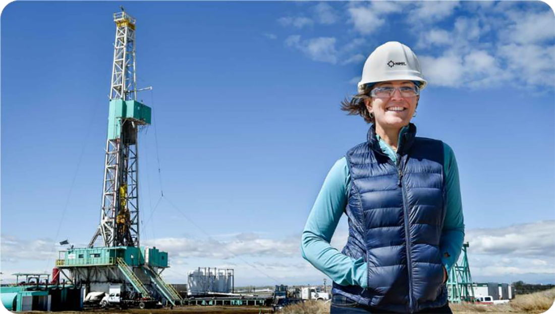 An NREL worker stands in front of a geothermal drill, wearing a hard hat, safety glasses, and a bright smile.