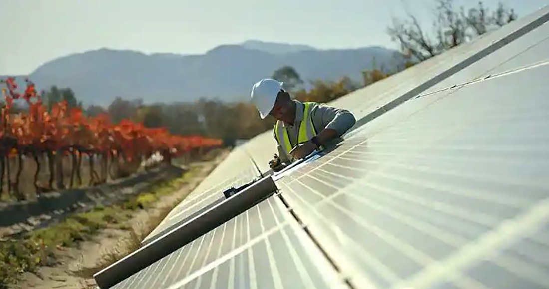 A man in a yellow vest and white hard hat works on solar panels outside with mountains in the background.