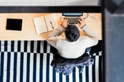 Person using a computer on a beige wooden desk, with a notepad, over a black and white rug