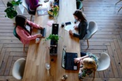 Four women sitting at a long wooden table with white chairs using computers and taking notes