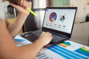 Person holding a highlighter marker viewing multicolor circle charts on a laptop on a desk along with more papers that include more graphs and charts