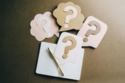 Cutout question mark symbols on paper speech bubbles are arranged above an open notebook with a pen on a brown surface.