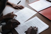 Two professionals review and point to contract documents on a table, with folders and paperwork spread across the workspace.