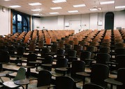 Empty lecture hall with tiered rows of wooden seats and attached desks under fluorescent lights and ceiling fans.