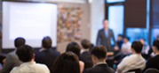 Professionals seated in a classroom listening to an instructor presenting at the front of the room.