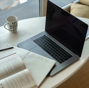 Person studies using a laptop, notebook and open book on a table, preparing for an online training or certification exam.