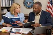 Two acquisition professionals review a Federal Acquisition Training Manual and certification study guide at a desk with a laptop.