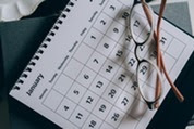 Eyeglasses rest on a calendar page beside a notebook, suggesting scheduling or planning.