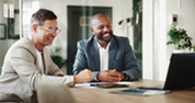 Two business professionals smile during a meeting at a conference table with a laptop, tablet and documents.
