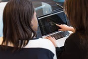 Two people collaborate at a laptop, with one pointing at the screen while the other looks on.
