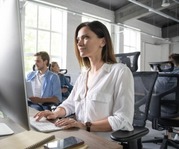 Woman works at a desktop computer in a bright office while coworkers work at nearby desks in the background.