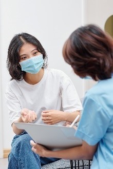 Image of a medical professional holding a chart and talking to a female patient wearing a face mask. 