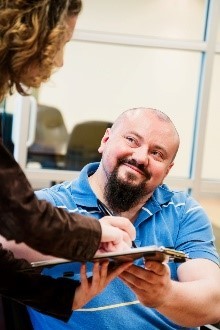 Smiling man signs a document on a clipboard in an office lobby.  
