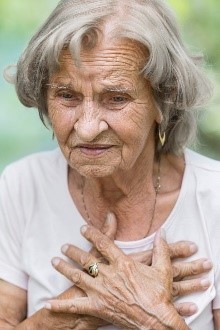 Elderly woman, seemingly with chest pain, holds her hands close to her heart. 