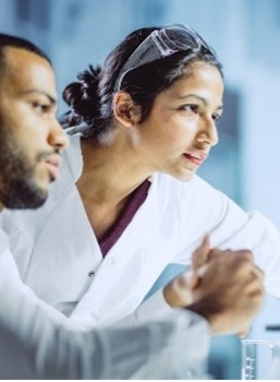 Photo of man and a woman wearing lab coats and looking into a computer screen.