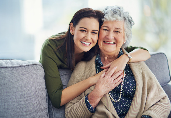 A young woman hugs her grandmother from behind while sitting on a couch