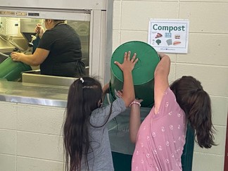 Two children dumping a green bucket of food waste into the large bin for composting