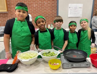 Five students wearing green aprons preparing to cook broccoli, radishes, and lettuce. 