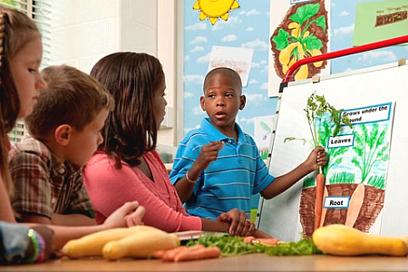 A young student holding a carrot and learning about growing vegetables with other students and a teacher. 