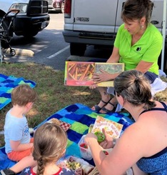 A family enjoying lunch on a blanket at the farmers' market while being read a book. 