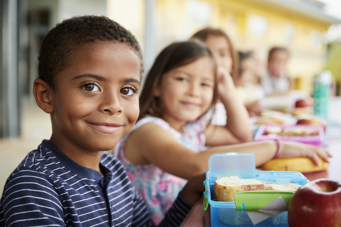 USDA recommends adding food safety items to your Back-to-School List. Image of children sitting at a cafeteria table enjoying their school lunch.