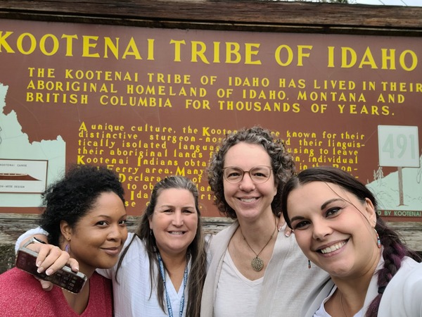 Four people standing in front of the Kooltenai Tribe in Idaho which is wood with yellow writing.