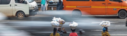 photo of street with vehicles passing by and women with baskets of goods on their heads