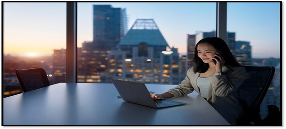 Woman on the cell phone and on the laptop in an office, window showing its dawn