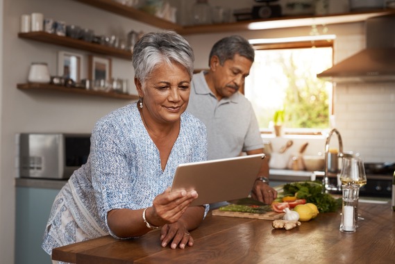 Older couple in the kitchen preparing food; Woman is viewing and holding a tablet.