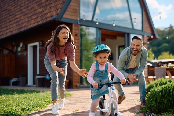 Parents hovering by child on a bicycle outside of a home on a brick pathway