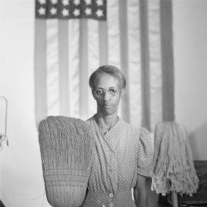 A woman stands holding a large broom with an American flag hanging in the background. Prints and Photographs Division