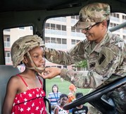 Photo of a service member helping a young girl in a red dress put on a military helmet. The girl is sitting in the cab of a truck.