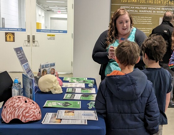 Woman next to event display table explains helmet safety to two boys