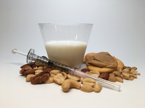 A syringe laying on mixed nuts in front of a glass of milk and wheat crackers with peanut butter spread.