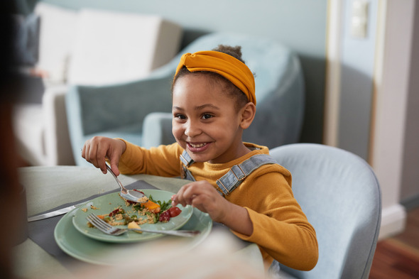 Young girl holding a fork sits at table with a plate of food in front of her and smiles at the camera. 