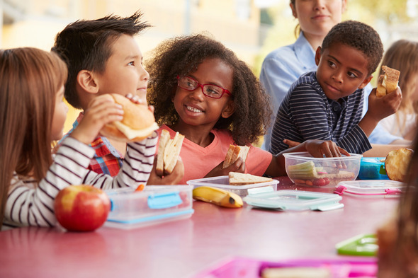 Three children look with interest at a fourth child while they eat together at a table.