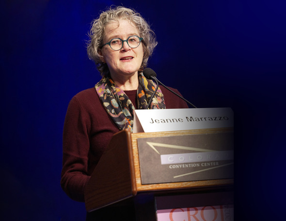 A person stands at a podium with "Jeanne Marrazzo" name card and "CROI" banner below