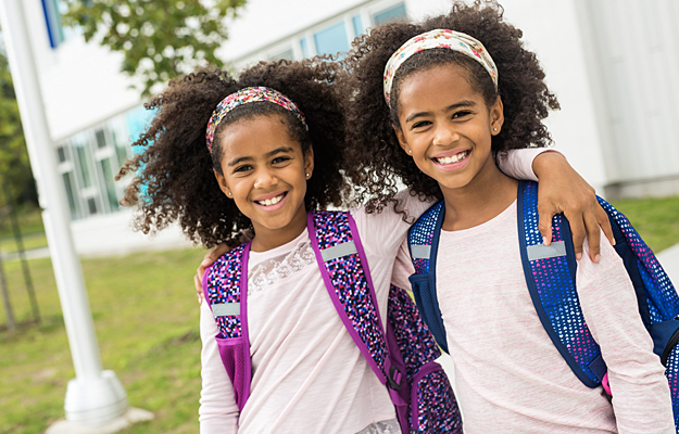 Twin sisters standing outside of school with backpacks on.