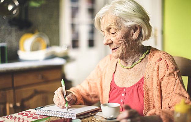 A senior woman in the kitchen writing in a notebook.