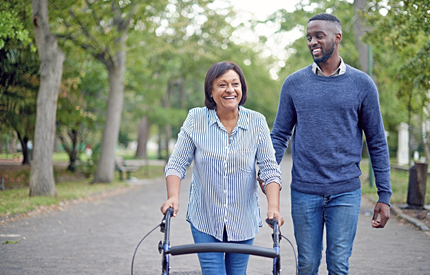 A mature woman using a walker while a young man assists her in the park.