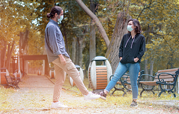 A man and woman wearing face masks and greeting each other by touching feet.