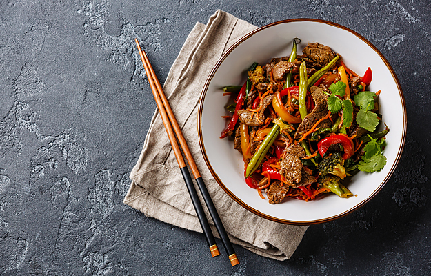 A bowl of beef stir fry with vegetables next to a napkin and pair of chopsticks.