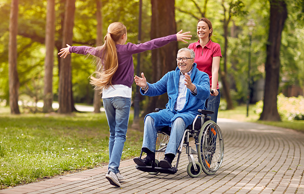 A caretaker pushing a senior man in a wheelchair towards a young girl with her arms out.