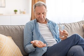 Older man sitting on a couch with a glass of water, looking at pills in his hand.