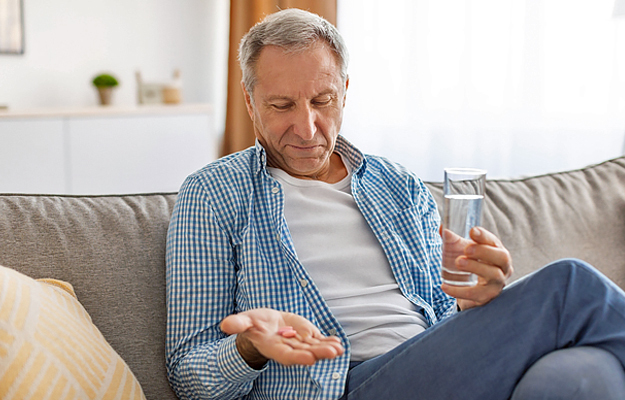 An older man sitting on a couch with a glass of water and looking at pills in his hand.