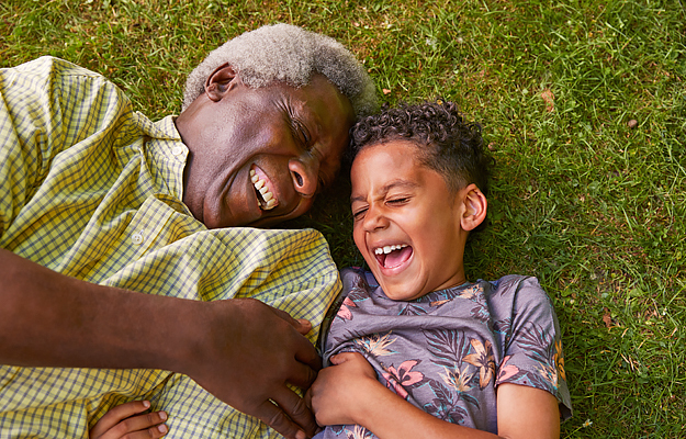 A senior man and his young grandson lying in the grass and laughing.