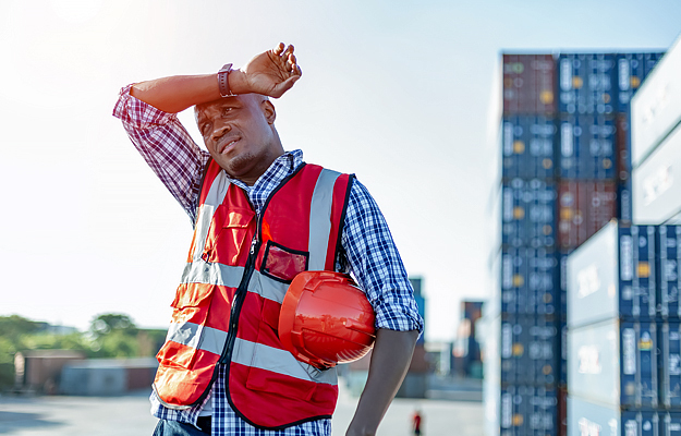 A male engineer in uniform on a sunny day holding a hard hat and wiping sweat from his brow.