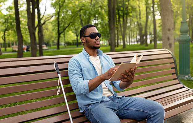 A vision impaired man sitting on a park bench and reading a braille book.